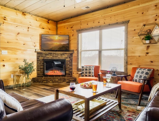 Log cabin living room with stone fireplace, electric insert, TV, orange chairs, brown leather seating, and large windows with blinds